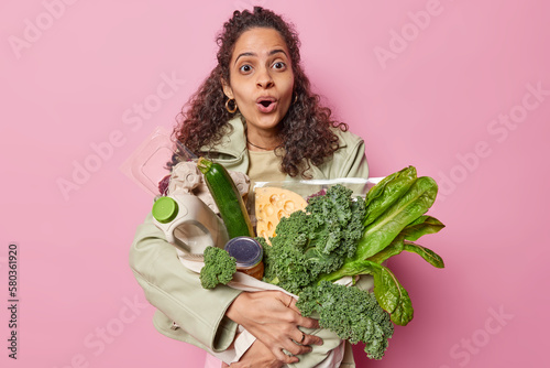 Surprised dark skinned teenage girl with purchase stares wondered at camera keeps mouth widely opened. Curly haired woman carries healthy products bought in supermarket poses against pink background