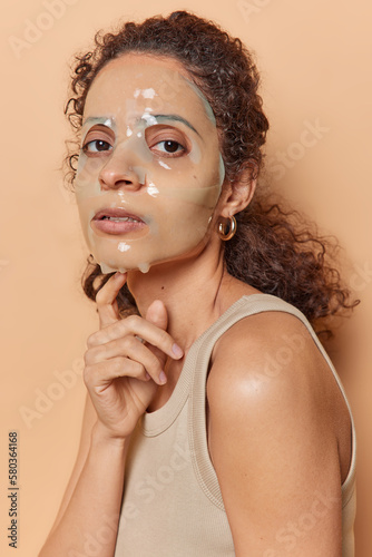 Vertical shot of curly haired woman applies beauty hydrogel sheet mask on face touches chin gently looks directly at camera dressed in casual t shirt isolated over brown background. Wellness concept