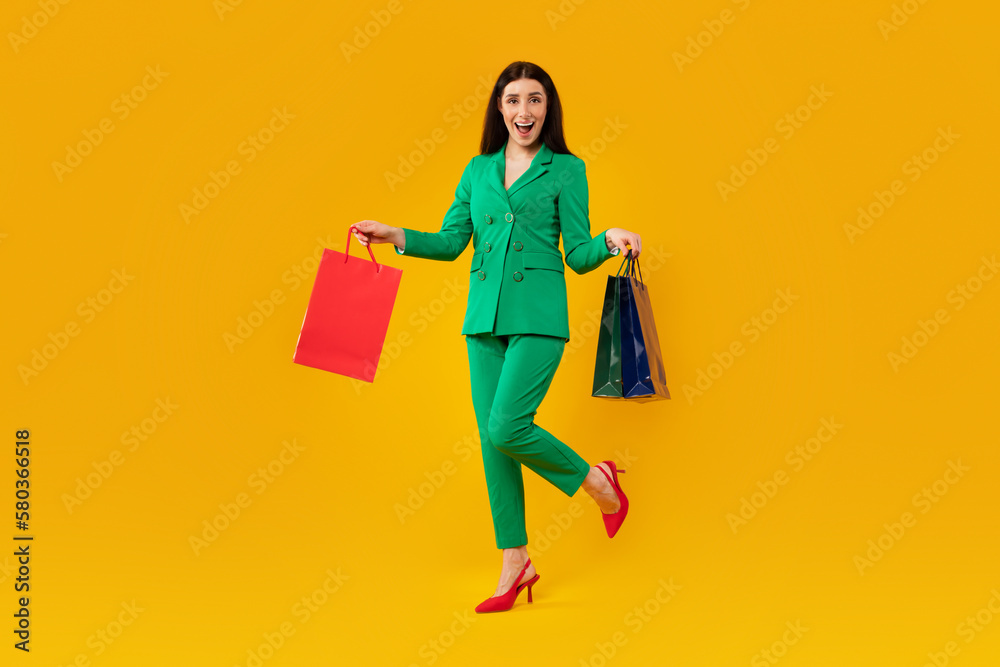 Excited woman in elegant outfit holding shopping bags and smiling, posing on yellow studio background, full length