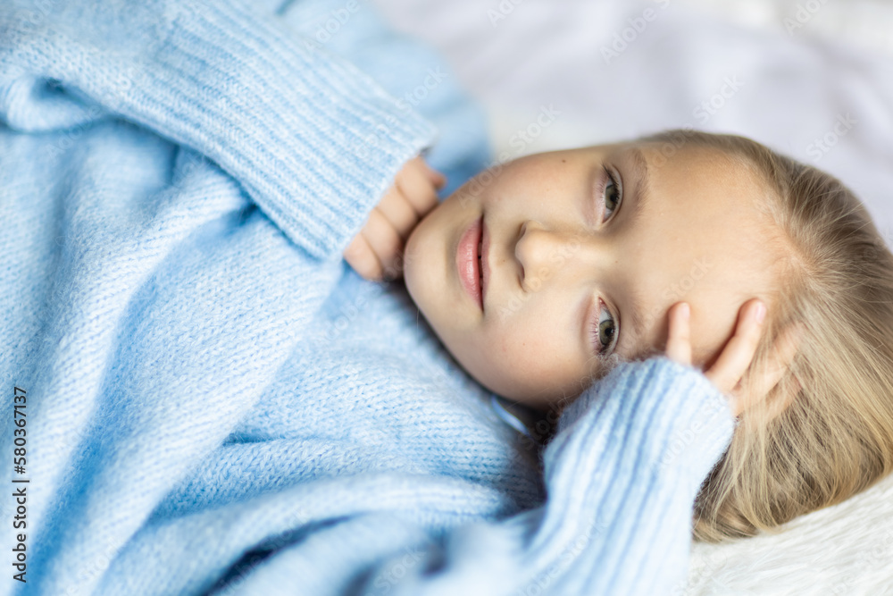 Cute little caucasian girl seven years old with blonde hair lying on bed at home in bedroom. Kid wearing stylish cozy knitted sweater blue color. Cute little caucasian girl seven years old with blonde hair lying on bed at home in bedroom. Kid wearing stylish cozy knitted sweater blue color.