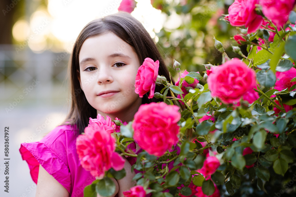 Smiling child girl 5-6 year old with rose flowers over blooming bushes in garden outdoor. Childhood. Spring season. Smiling child girl 5-6 year old with rose flowers over blooming bushes in garden outdoor. Childhood. Spring season.