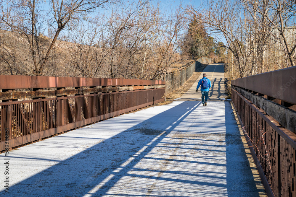 older man with a backpack is walking on a biking trail and footbridge covered by frost, winter morning in Fort Collins, Colorado