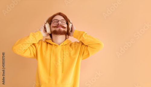 A stylish young man with red hair and an outrageous red mustache  enjoys music playing in headphones.  The man raised his head up and holds headphones near his ears with both hands and smiles.