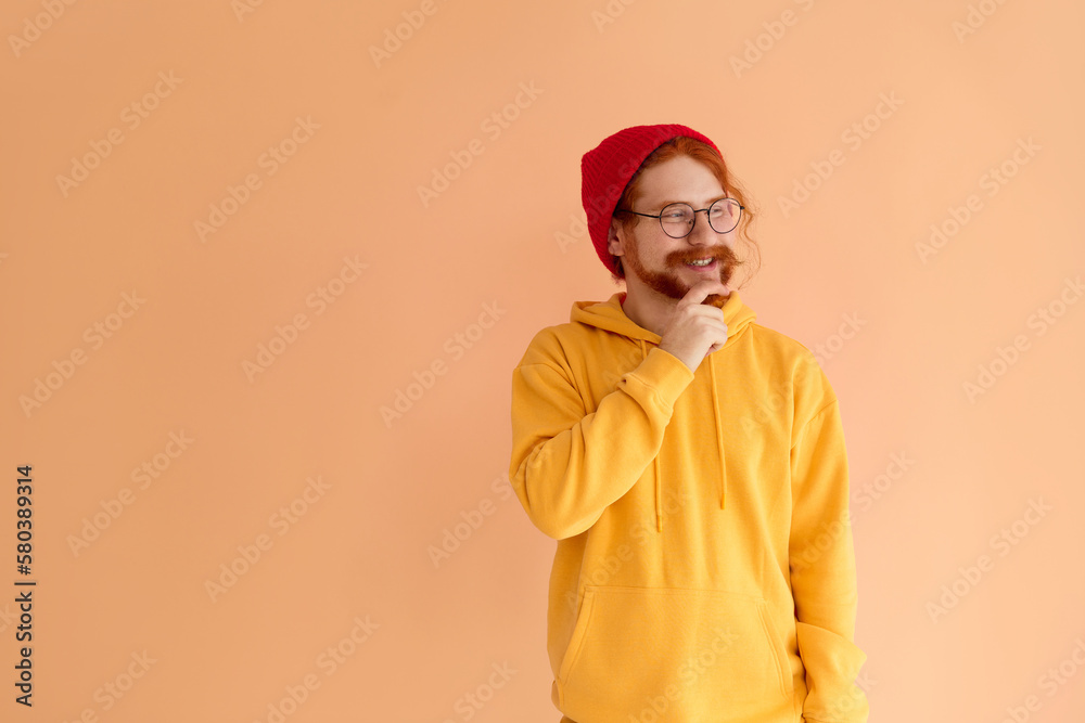 A stylish young man in glasses wearing a red cap and a yellow hoodie looks thoughtfully to the side and smiles.Copy space.