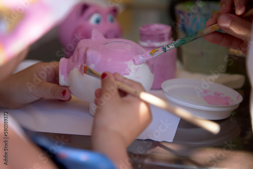 The child's hand holds a brush and paints a clay piggy bank in the shape of a pig