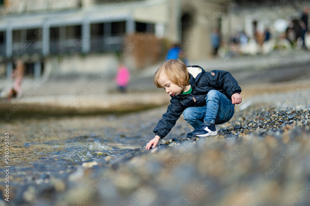 Cute toddler boy playing with pebbles in the harbor of Varenna, one of the most picturesque towns on the shore of Lake Como. Varenna, Italy. Cute toddler boy playing with pebbles in the harbor of Varenna, one of the most picturesque towns on the shore of Lake Como. Varenna, Italy.