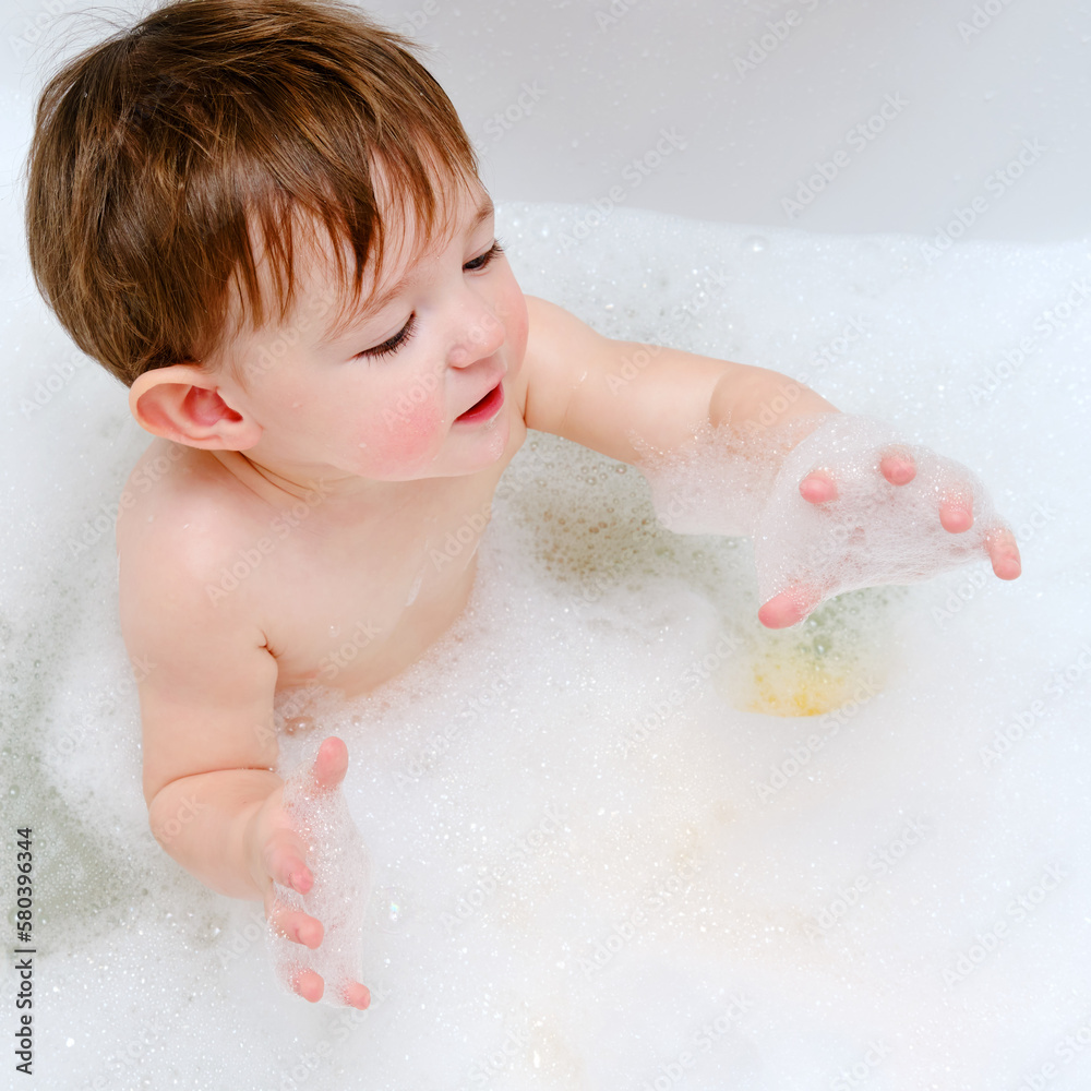 A happy child is playing with soap bubbles while sitting in a white home bathtub. Smiling toddler baby boy bathes in the bathroom. Kid aged one year six months A happy child is playing with soap bubbles while sitting in a white home bathtub. Smiling toddler baby boy bathes in the bathroom. Kid aged one year six months