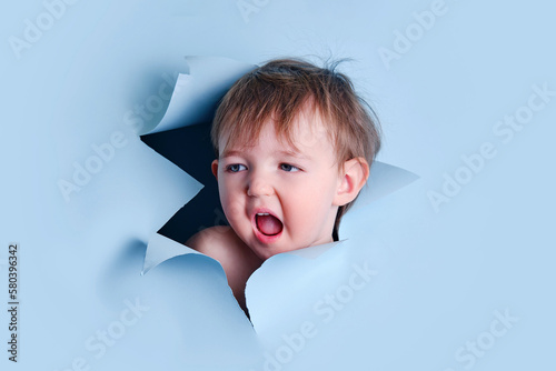 A yawning baby in a hole on a paper blue background. Torn child's head studio background, copy space. Kid aged one year six months photo