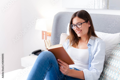 Young woman reading book at home. Happy girl in spectacles sitting in bed. Student studying in comfortable bedroom. Leisure  mental health  relaxing. Lifestyle moment