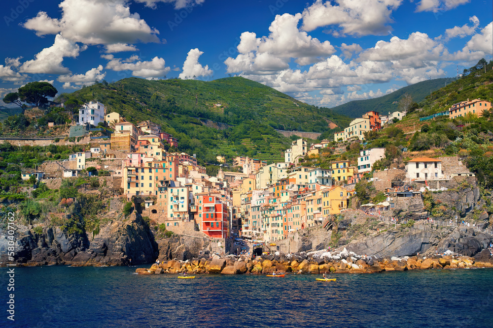Picturesque village of Riomaggiore against a blue sky with white clouds. View from the sea side of the colourful houses, green terraced vineyards, tourists enjoying holidays. Cinque Terre, Italy.