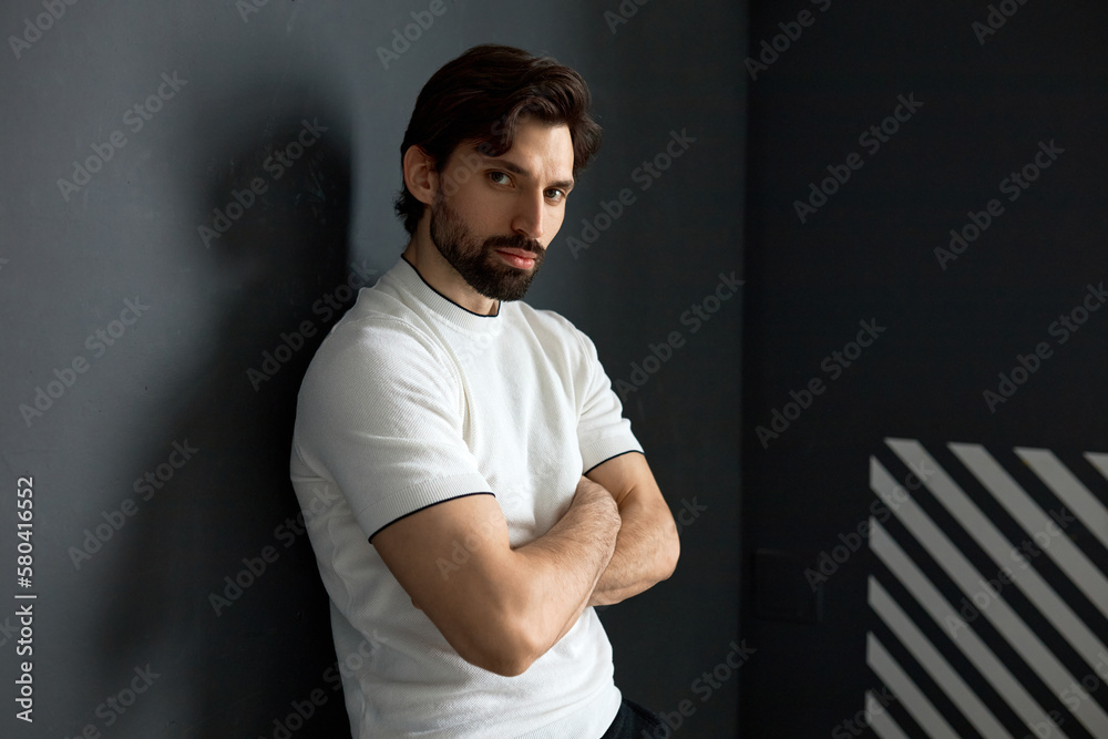 Strong manly confident millennia bearded brunet guy in white sport t-shirt at gym, attractive athletic male fitness trainer posing against black wall of health club with folded arms Strong manly confident millennia bearded brunet guy in white sport t-shirt at gym, attractive athletic male fitness trainer posing against black wall of health club with folded arms