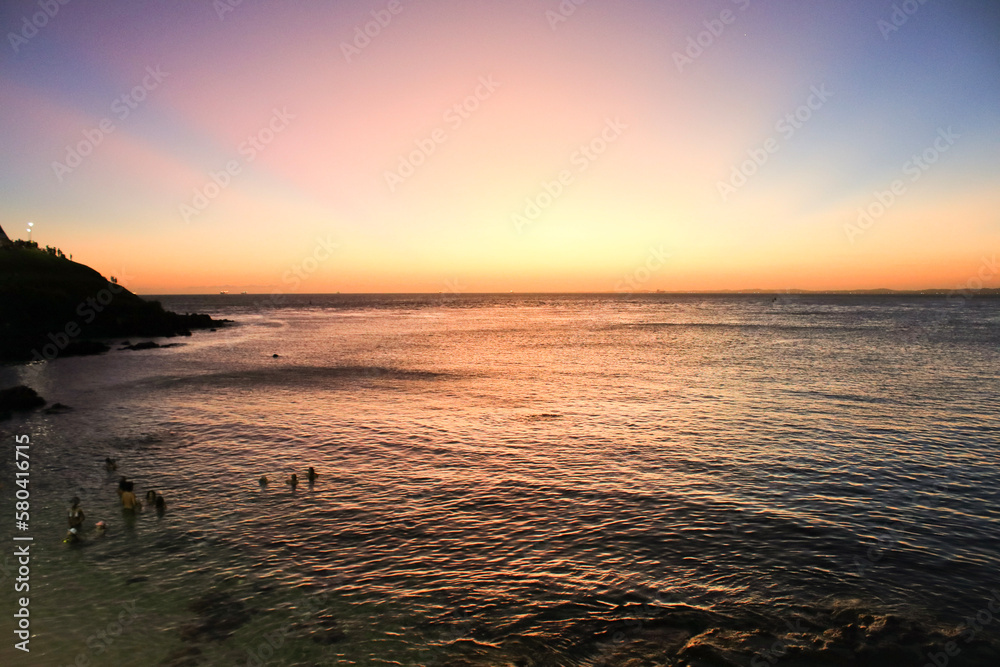 People swimming and enjoying the wonderful sunset on the beach with sand and stones and the blue sky with sun rays in yellow, orange and red colors