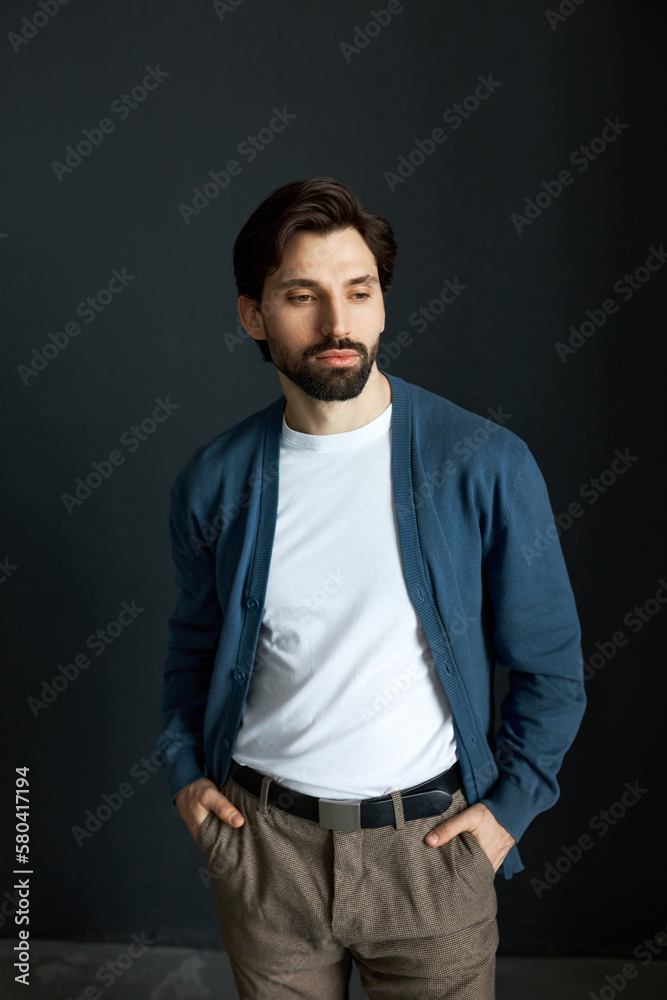 Portrait of handsome brutal man teacher in cardigan and stylish pants standing against black studio background with hands in pockets, looking down, preparing his mind for productive work day