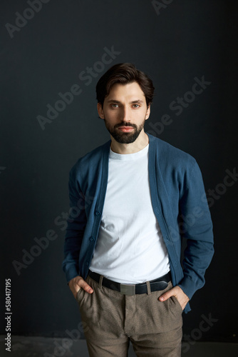 Charming male fashion model in blue cardigan and plaid brown pants posing on black studio background with hands in pocket, looking at camera with confident facial expression. Fashionable menswear © Anatoliy Karlyuk