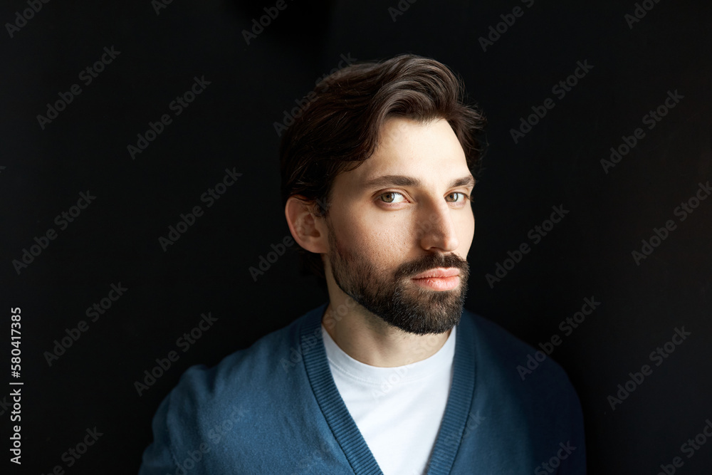 Head portrait of charming brutal millennial brunet guy looking at camera with suspicious uncertain facial expression, posing against black background with shadow falling on his shoulder