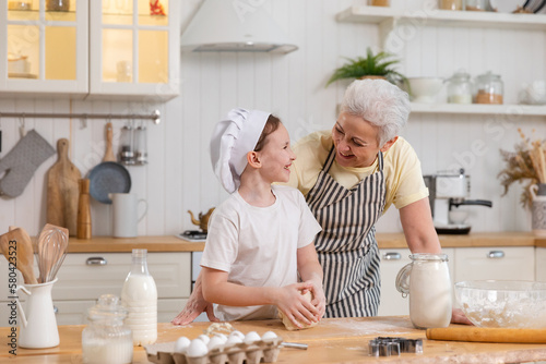 Happy family in kitchen. Grandmother and granddaughter child cook in kitchen together. Grandma teaching kid girl knead dough bake cookies. Household teamwork helping family generations concept.