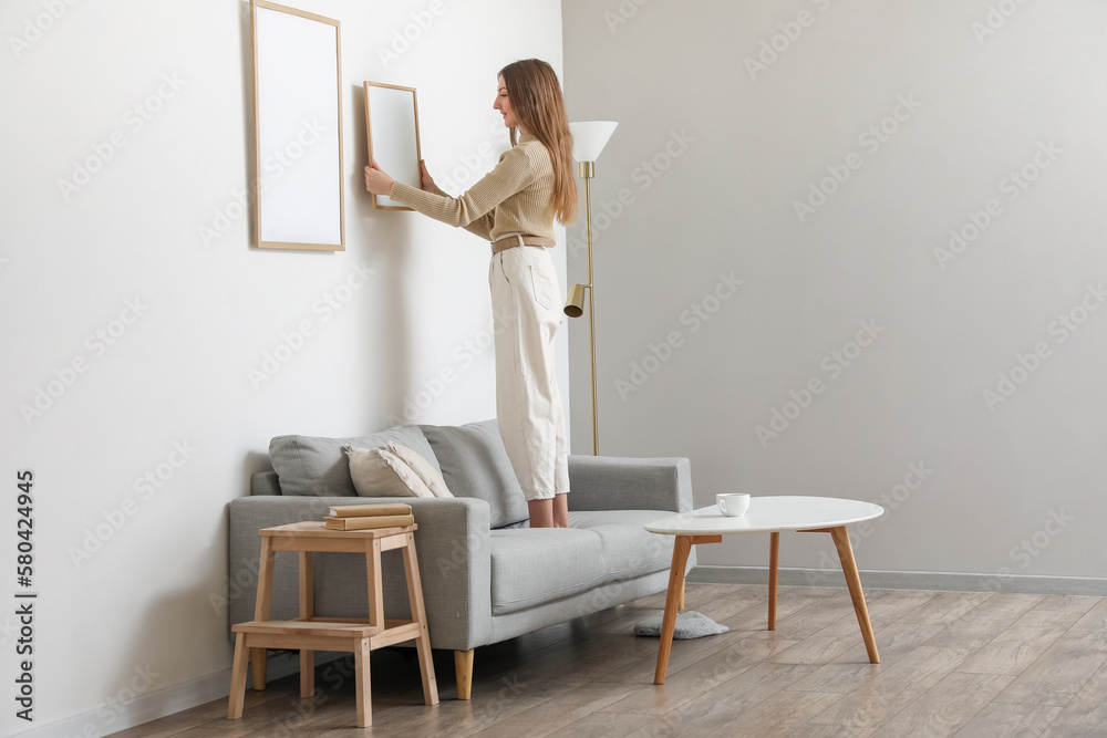 Young woman hanging blank frame on light wall at home - obrazy, fototapety, plakaty 
