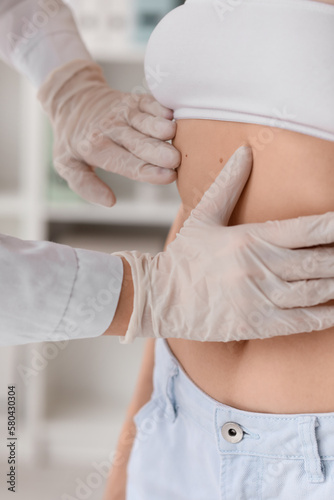 Dermatologist examining moles on young woman's belly in clinic, closeup