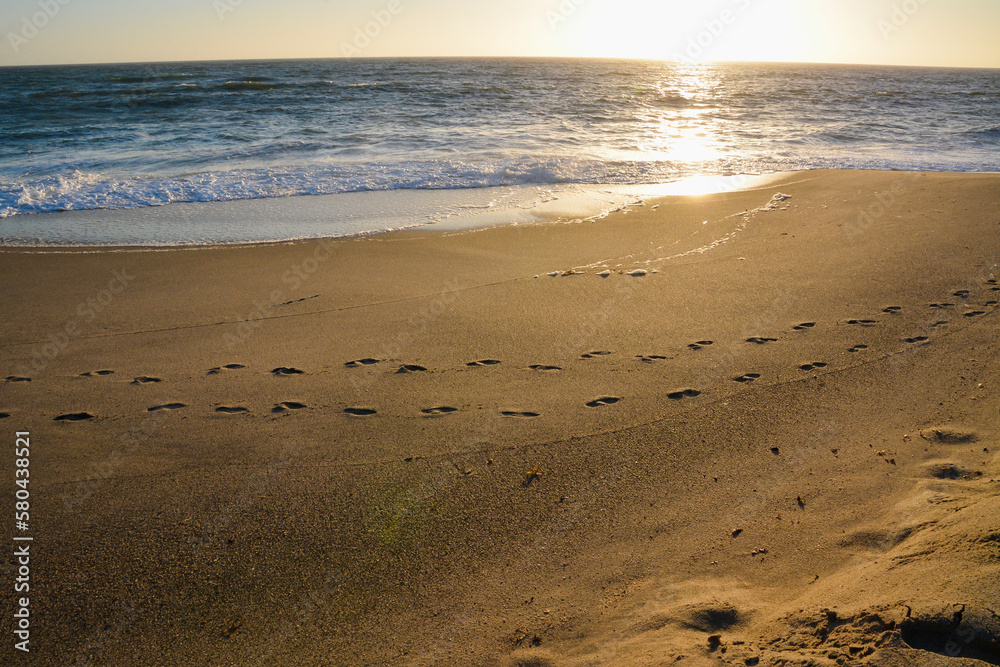 Footprints on a sea beach parallel to the shore. Waves splash on the shore. The sun reflects in the water Footprints on a sea beach parallel to the shore. Waves splash on the shore. The sun reflects in the water