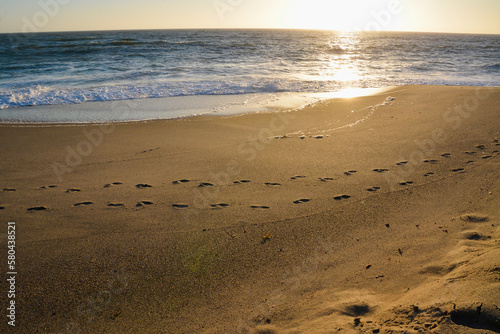 Footprints on a sea beach parallel to the shore. Waves splash on the shore. The sun reflects in the water