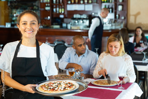Young cheerful hispanic woman pizzeria owner inviting to taste delicious pizza in cozy atmosphere