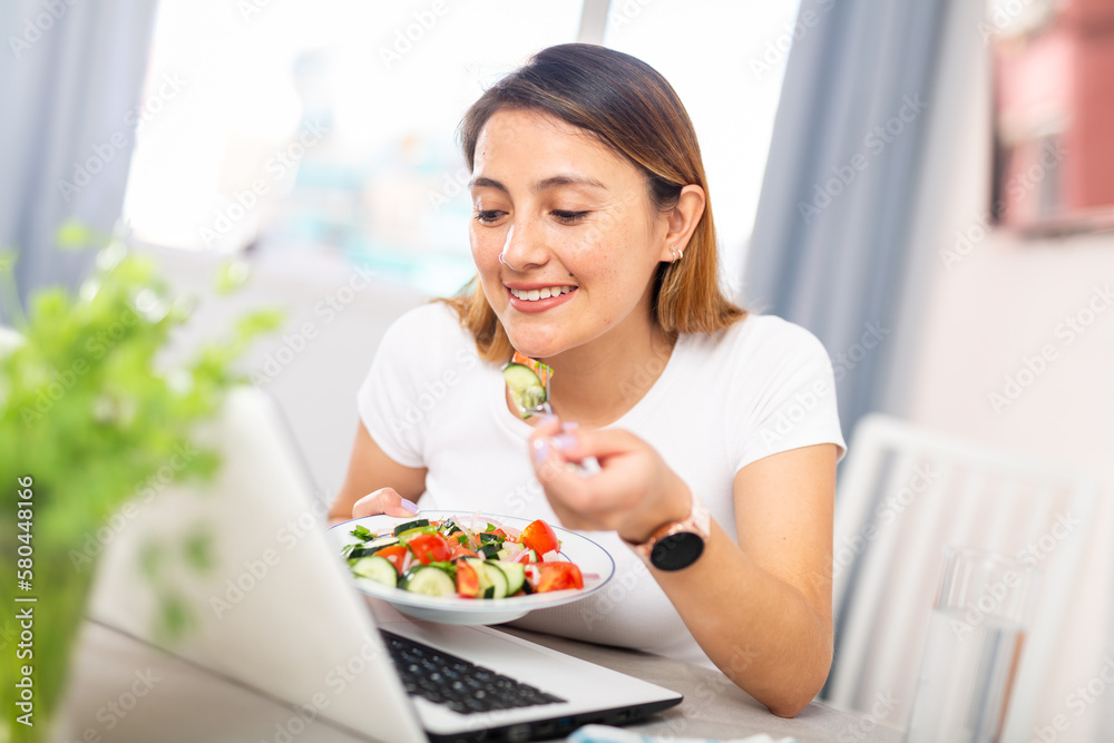 Positive young latino woman eating salad and chatting online using laptop