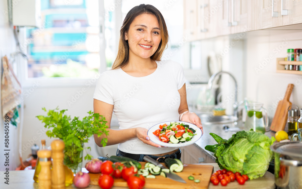 Positive colombian housewife before eating vegetable salad from plato at kitchen