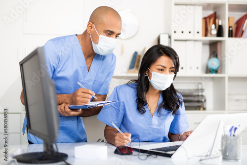 Professional doctor in surgical mask working in office with female assistant, filling up medical forms in laptop