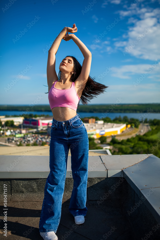 Beautiful sexy slim brunette caucasian girl in pink top and blue jeans standing on the roof attractively posing with softly blurred sky background