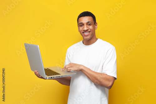 young african american guy in white t-shirt uses laptop and types on yellow isolated background, man holds computer