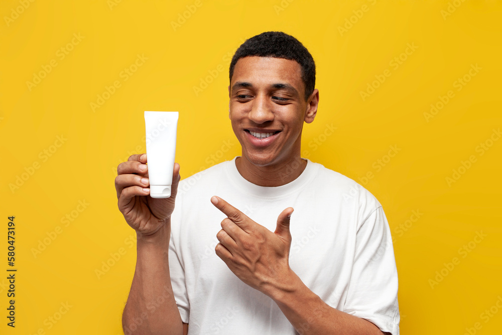 young african american guy in white t-shirt shows tube of cream on yellow isolated background young african american guy in white t-shirt shows tube of cream on yellow isolated background