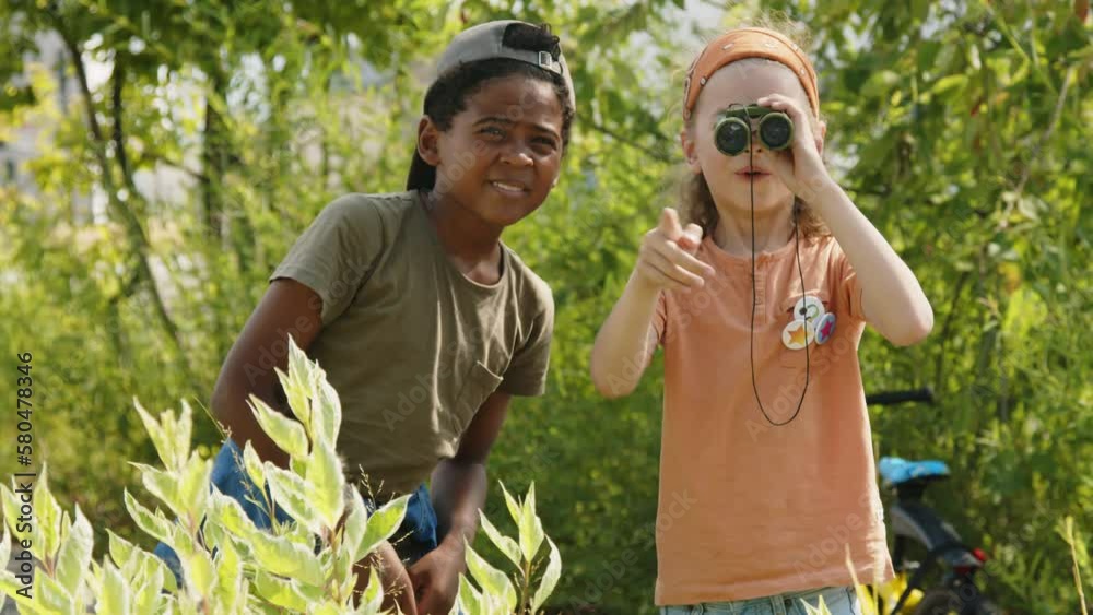Medium shot of young African American and Caucasian boys playing outdoors in summer, standing in green bushes, looking through binoculars, chatting and pointing at something far away