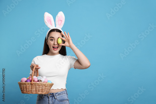 Happy woman in bunny ears headband holding wicker basket of painted Easter eggs on turquoise background. Space for text