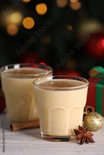 Tasty eggnog, cinnamon, anise and Christmas decorations on white wooden table against blurred lights