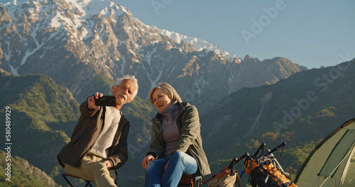 Senior cacasian couple having a rest on top of a mountain, taking a picture or having video chat on smartphone, travelling together after retirement - pension, recreational pursuit, tourism concept  photo