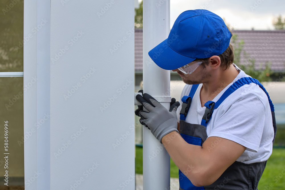 Man in a blue cap and jumpsuit mounts a new metal gutter system on the house. New white downspout is installed by a professional builder