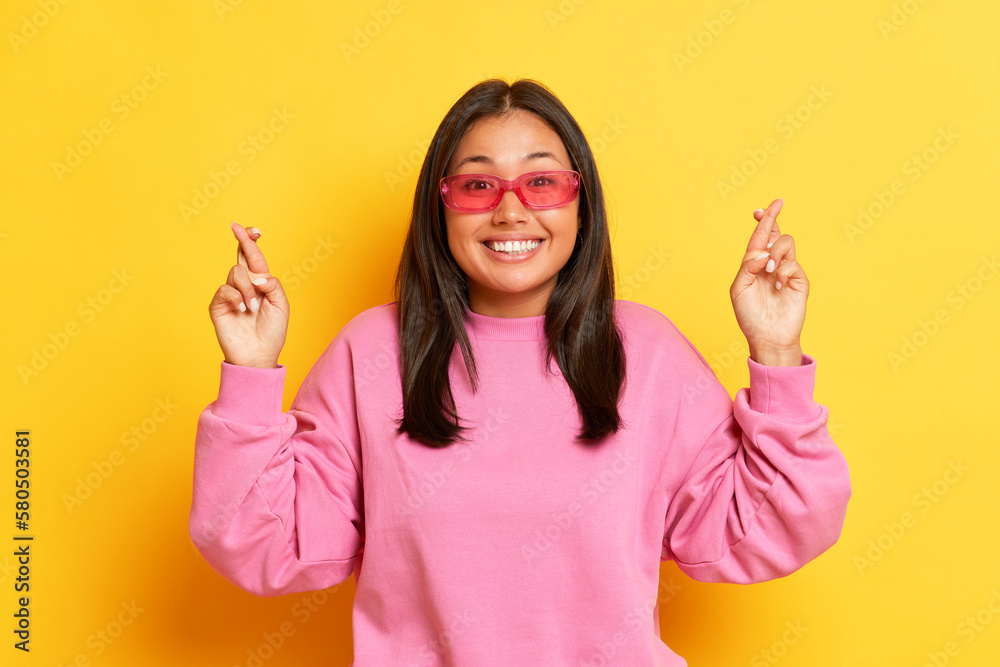 Pretty smiling girl in pink sunglasses standing on yellow background, student crosses her fingers for good luck before the exam, copy space, high quality photo Pretty smiling girl in pink sunglasses standing on yellow background, student crosses her fingers for good luck before the exam, copy space, high quality photo
