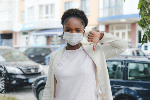 Discontent African American woman wearing medical mask showing dislike with thumbs down, rejection concept. Concept of coronavirus, quarantine and social