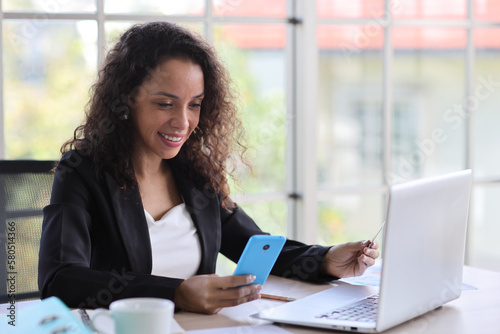 Attractive professional latin female employee worker sitting, using laptop computer with paperwork at home workplace. Businesswoman talking on smart mobile phone and communicate with client.