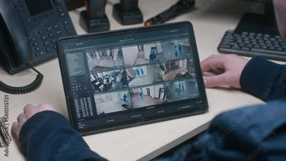Security officer controls CCTV cameras in coworking office, uses digital tablet with surveillance cameras playback on screen. High tech security with facial recognition. Monitoring and social safety.