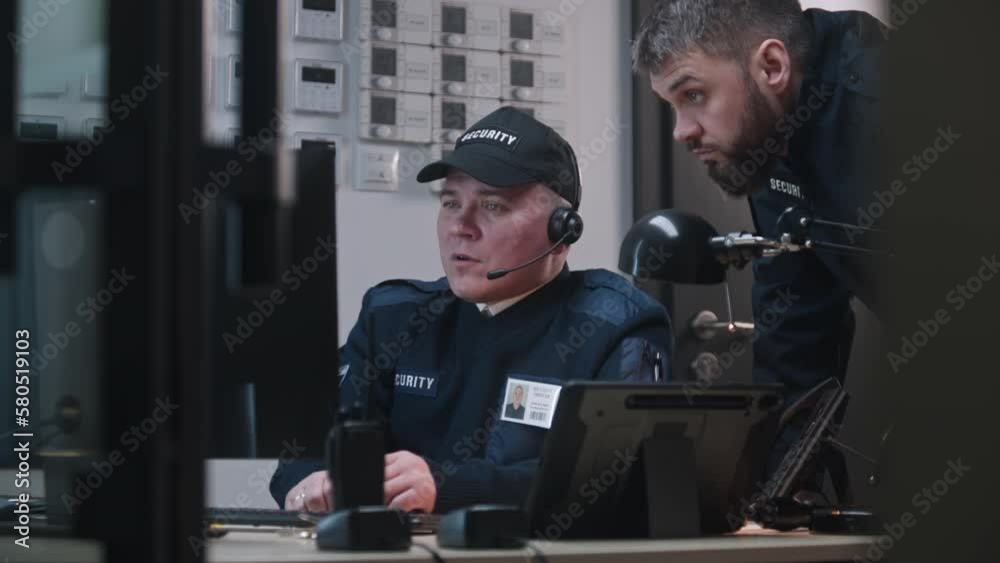 Man comes to colleague, looks view from security cameras displayed on computer and tablet monitors and talks by walkie talkie. Security officer sits at workplace and typing keyboard. CCTV technology. Man comes to colleague, looks view from security cameras displayed on computer and tablet monitors and talks by walkie talkie. Security officer sits at workplace and typing keyboard. CCTV technology.