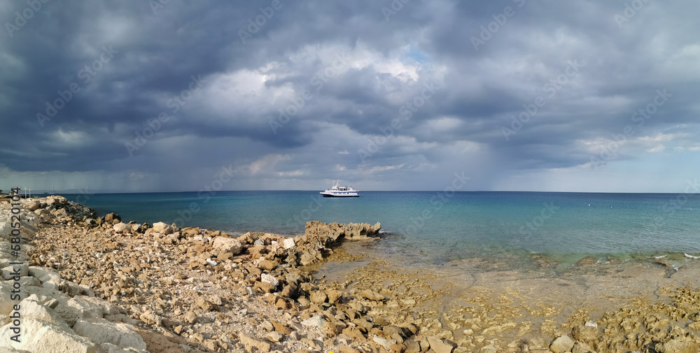 The stone coast of the Mediterranean Sea, a white ship in the sea and the largest sailing yacht in the world against a dramatic sky.