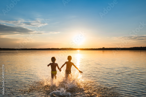 Happy children runs splashing water on beach towards the sun. Back view, wide angle shooting.