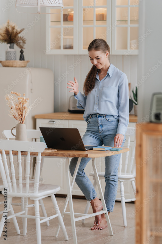 Successful young student girl in jeans and shirt standing at kitchen table with laptop talks via internet with parents. Cheerful woman in casual makes video call home enjoys weekend. Domestic leisure. Successful young student girl in jeans and shirt standing at kitchen table with laptop talks via internet with parents. Cheerful woman in casual makes video call home enjoys weekend. Domestic leisure.
