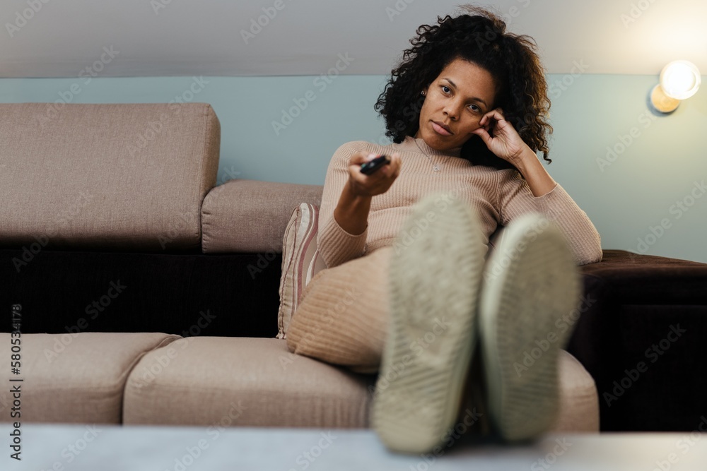 Afro american woman sitting on a couch at home, watching TV, holding remote control Afro american woman sitting on a couch at home, watching TV, holding remote control