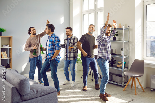 Happy male friends having party  dancing and drinking beer  having fun together in the living room at home. Group of brutal friendly  positive men in casual clothes enjoying spending time indoors.