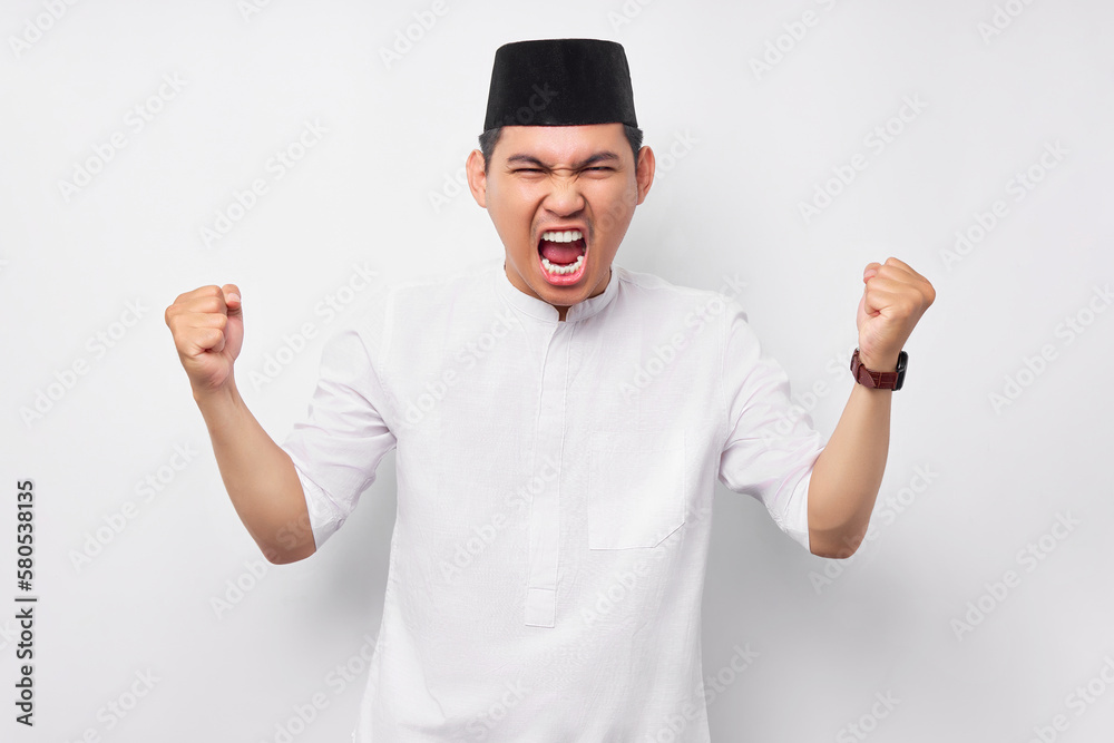 Fototapeta premium Overjoyed young Asian Muslim man screaming and raising a hand isolated on white background. People religious Islamic lifestyle concept