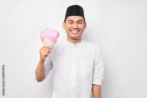 Portrait of smiling handsome young Asian Muslim man holding cash money rupiah banknotes isolated on white background. People religious Islamic lifestyle concept