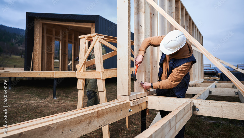 Man worker building wooden frame house on pile foundation. Carpenter hammering nail into wooden truss, using hammer. Carpentry concept. Man worker building wooden frame house on pile foundation. Carpenter hammering nail into wooden truss, using hammer. Carpentry concept.