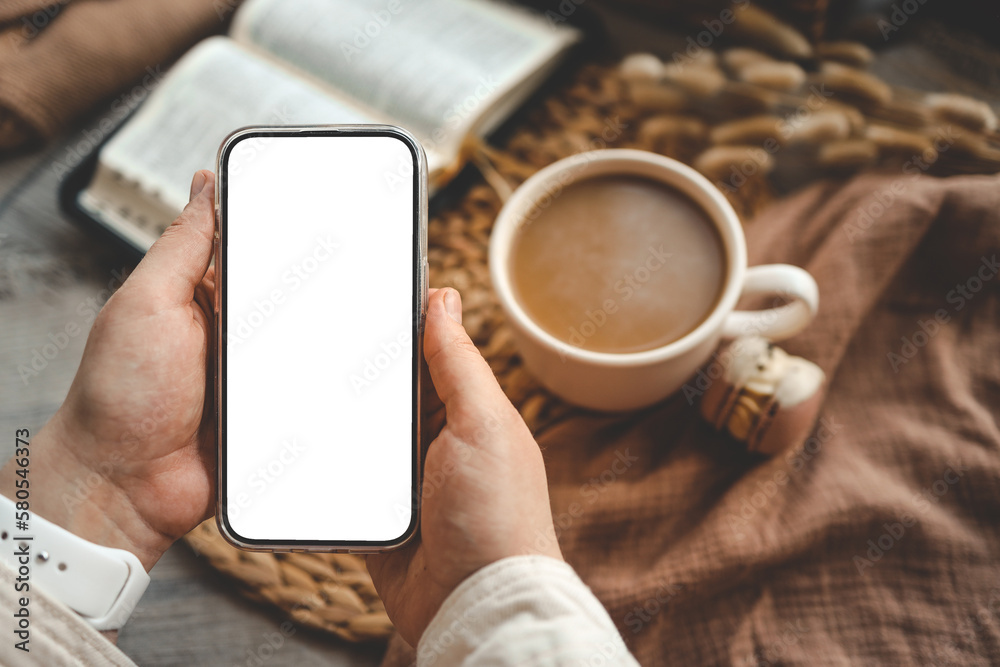 Hands holding a phone with an isolated screen, seen from above, with an open Bible, a coffee cup, and macarons in a home interior Hands holding a phone with an isolated screen, seen from above, with an open Bible, a coffee cup, and macarons in a home interior